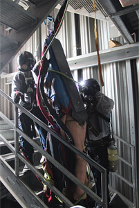 2 Firefighters guiding a training manikin down a narrow dark stairwell.