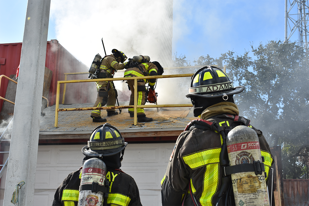 2 firefighters watch 2 more attempt a roof entry of a burning garage during a scenario. 